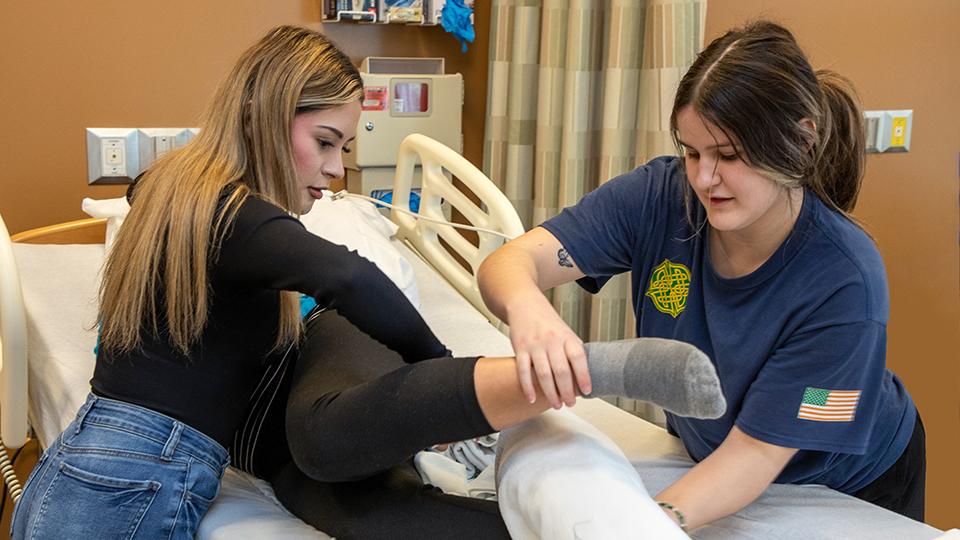 Two students assist a patient in a hospital bed.